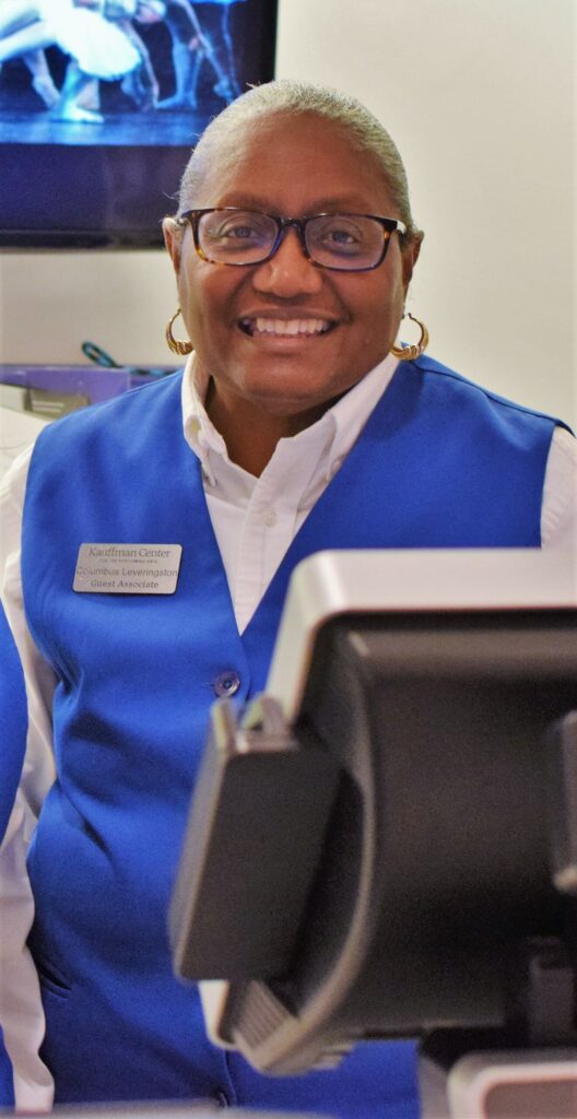 A person wearing a blue vest and glasses is smiling at the camera. They have a name tag on their vest. A computer monitor is visible in the foreground.