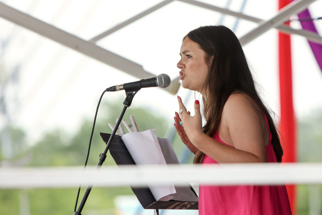A person in a pink dress speaks or sings into a microphone on stage, with sheets of paper on a music stand in front of them.