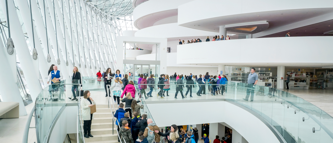 People walking and standing on the upper level of a modern indoor public space with large windows and white architectural elements, reminiscent of scenes from A Guide to the Kauffman Center.