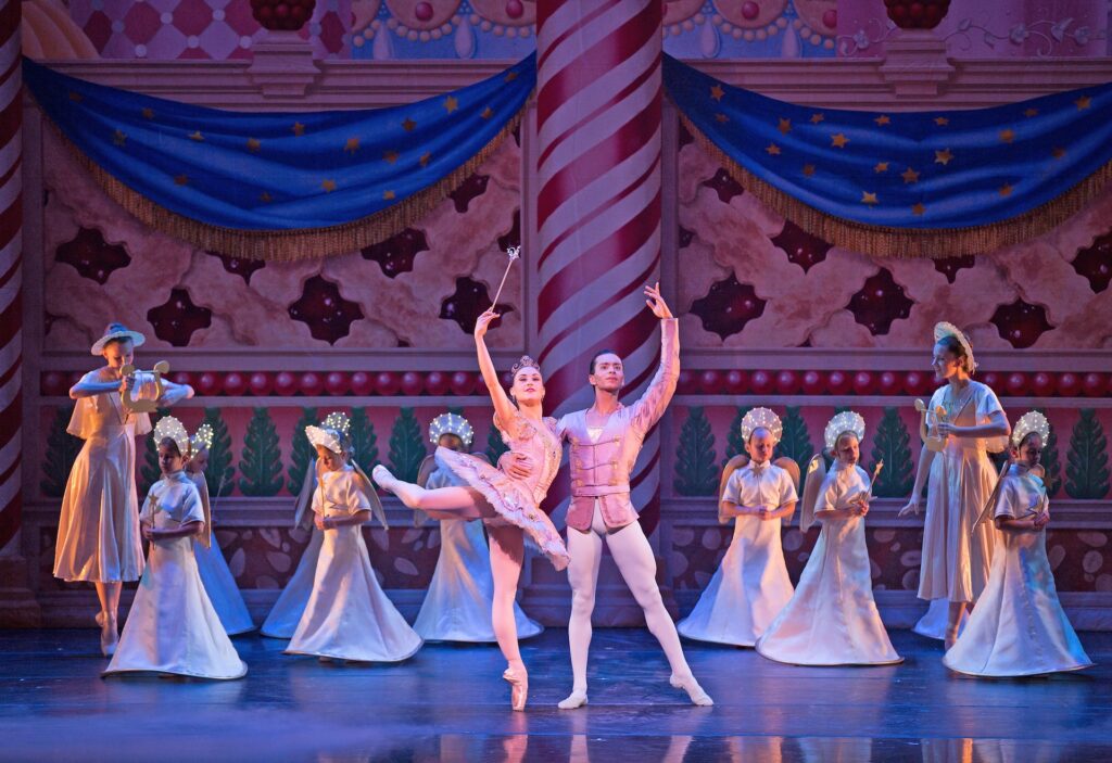 Ballet dancers perform on stage in ornate costumes, with a pink-decorated backdrop. A female dancer balances on one leg, while a male dancer stands beside her. This captivating scene feels like a highlight from the Performing Arts Sampler, with other dancers gracefully poised in the background.