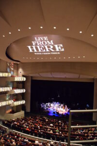 View of a theater interior with an audience watching a live performance. The stage is lit up, and "Live From Here with Chris Thile" is projected on the ceiling above the stage.