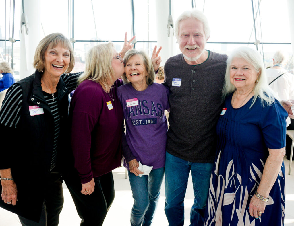 Five people standing together indoors, smiling and posing for the photo. One person playfully makes a peace sign behind another's head. They have name tags and are dressed casually.