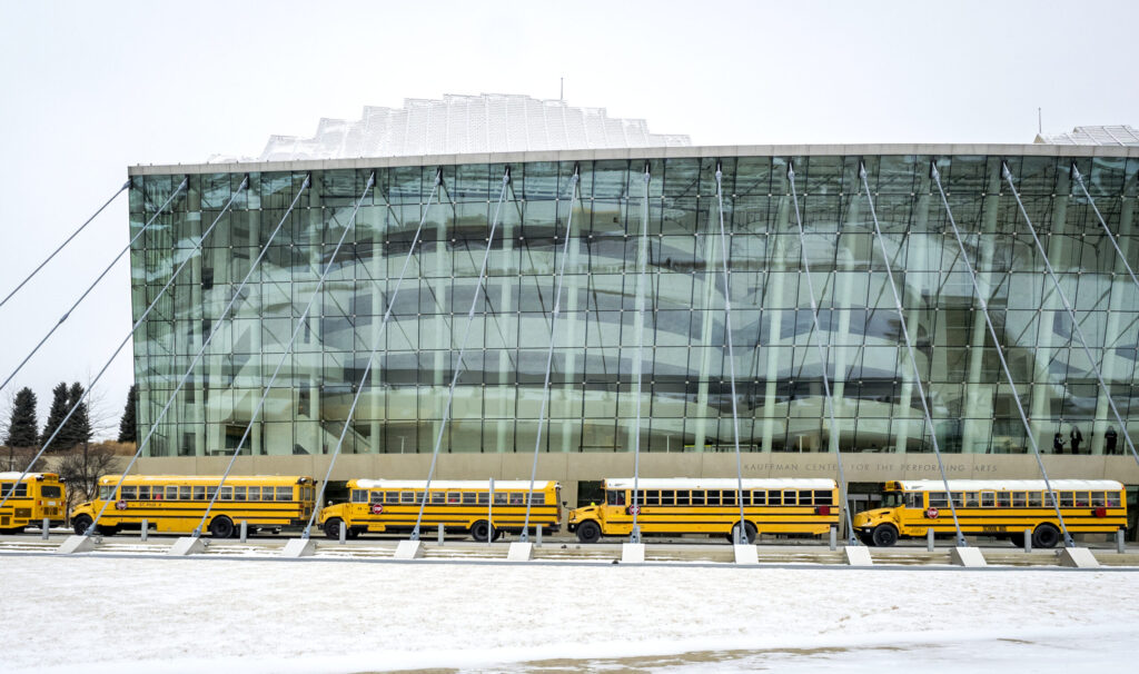 A row of yellow school buses is parked in front of a glass-fronted modern building on a snowy day.