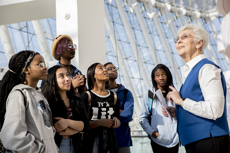 A group of students attentively listens to an elder woman, who is presumably a guide, giving a tour in the modern, glass-roofed building of the Kauffman Center.