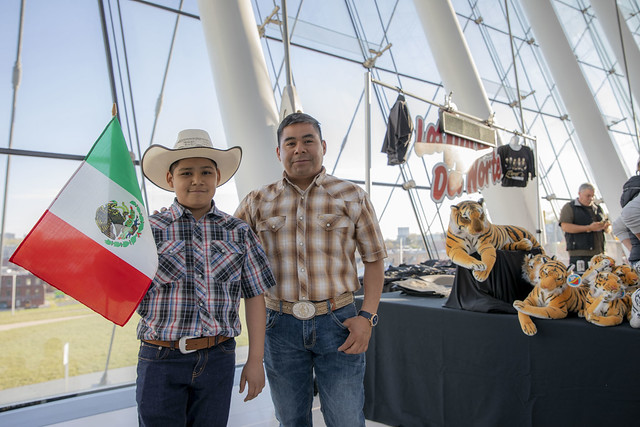 A boy holding a Mexican flag stands next to an adult man in a plaid shirt and cowboy hat. They are at an indoor event with stuffed tiger toys displayed on a table in the background.