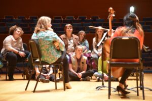 A group of people, including children and adults, are seated in a semi-circle listening to a cellist perform in a large auditorium.
