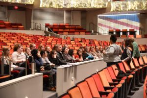 A group of people sit in a theater's orange seats, listening to a few individuals standing in front of them. The theater has a balcony and modern design elements.