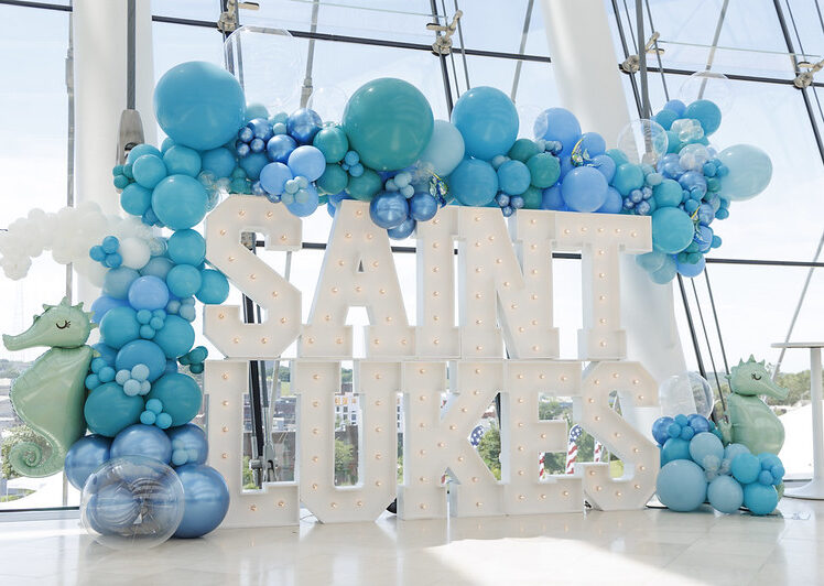 A large display with illuminated letters spelling "Saint Luke's," adorned with blue and white balloons and seahorse decorations, set in a modern, glass-walled venue.