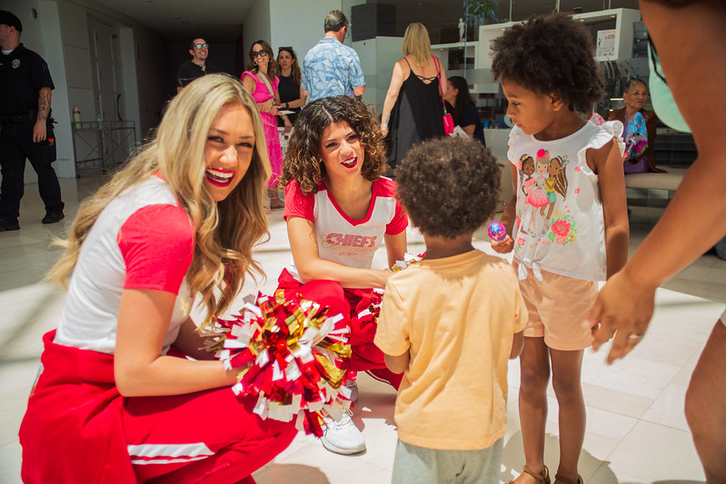 Two chiefs cheerleaders in red uniforms kneel and interact with three young children in a brightly lit indoor setting, while onlookers observe in the background. In the News recently, such heartfelt moments of innocence and connection continue to captivate audiences everywhere.