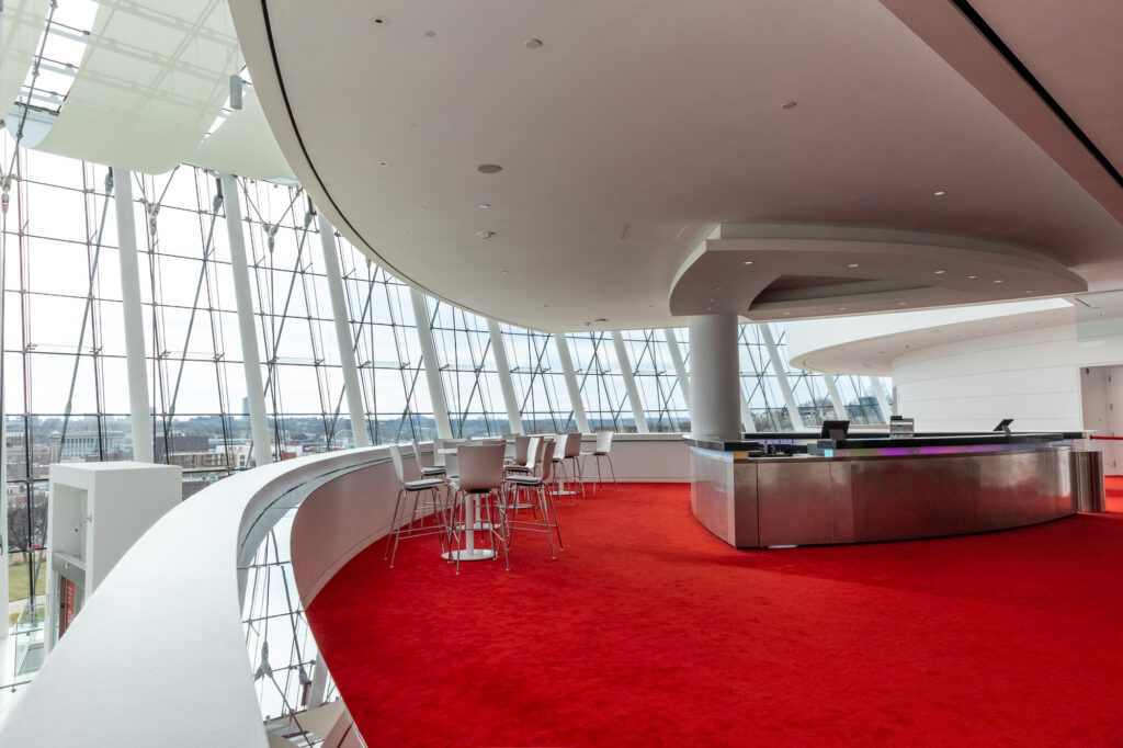 Modern interior space with red carpeting, high glass walls, and a curved white ceiling. A row of chairs and tables is set near the windows, and a metallic counter is on the right side.