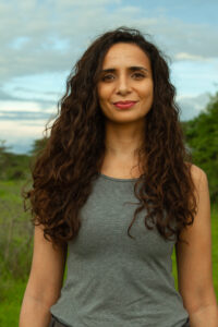 A woman with long, curly hair wearing a gray tank top stands outdoors with greenery and a cloudy sky in the background.