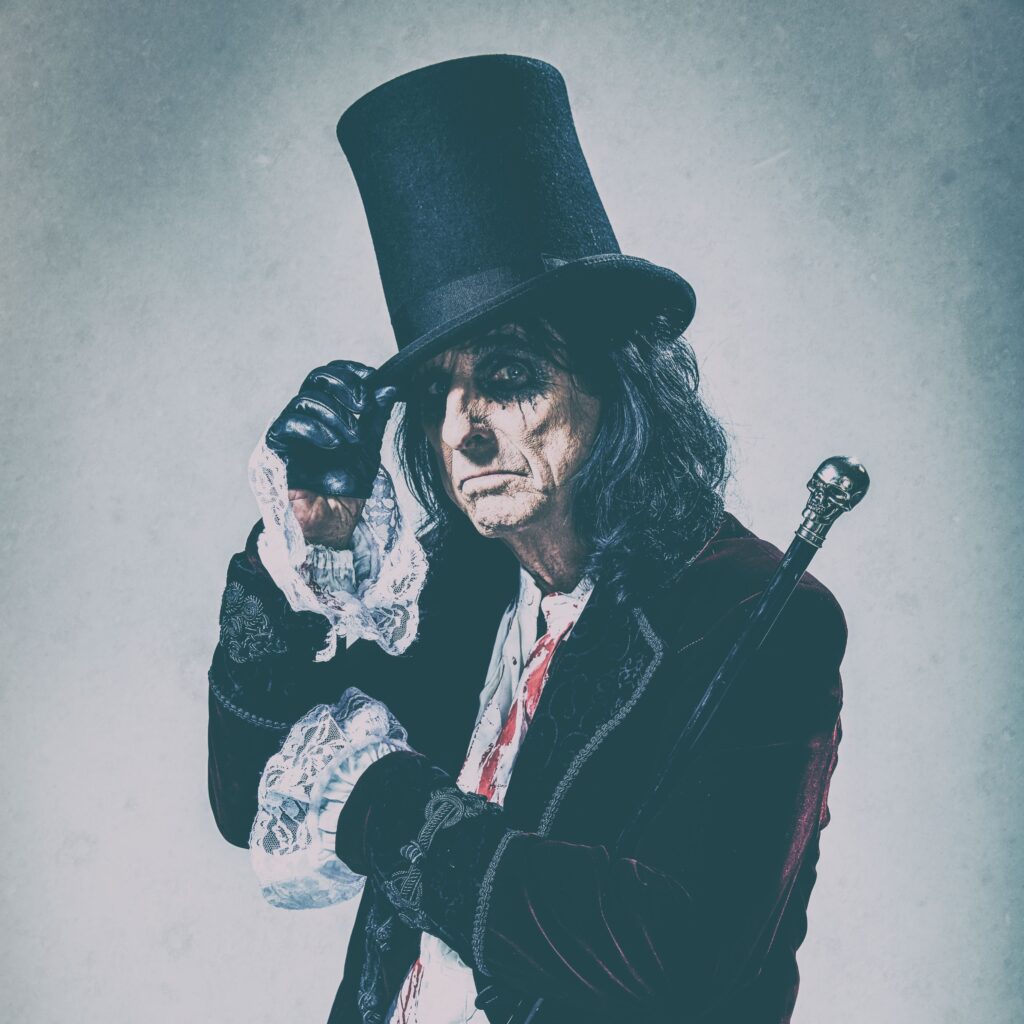 Person in a vintage-style outfit with a tall black hat and dark coat, holding a cane, and adjusting the hat with one hand while looking at the camera.