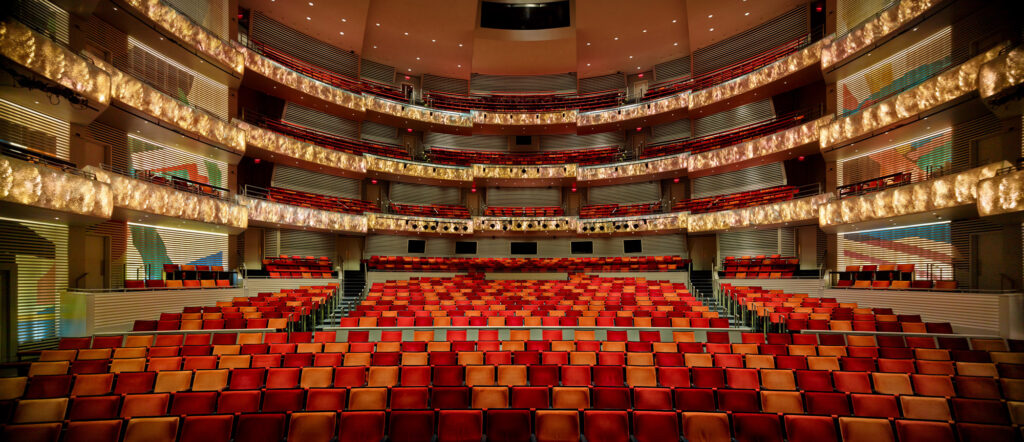 An empty, modern theater with red and beige seats arranged in a semi-circular pattern across multiple tiers.