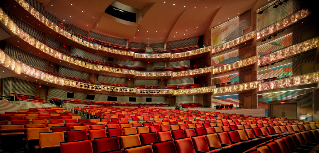 A spacious, multi-tiered theater with rows of red and orange seats, illuminated stage lighting, and decorative interior balconies.