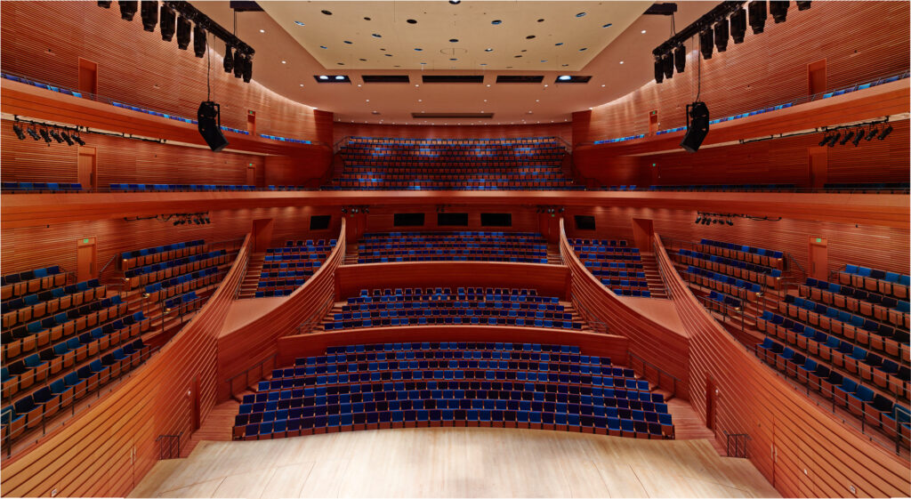 A view of an empty modern concert hall with multiple tiers of seating, wooden interior walls, and a well-lit stage. The seats are blue with wooden backs, and there are spotlights mounted above.