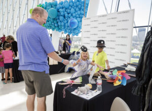 An adult and two children are at a craft table with masks and various items. A backdrop with "Lyric Opera" logos and blue balloon decorations are in the background. They are inside a modern building.