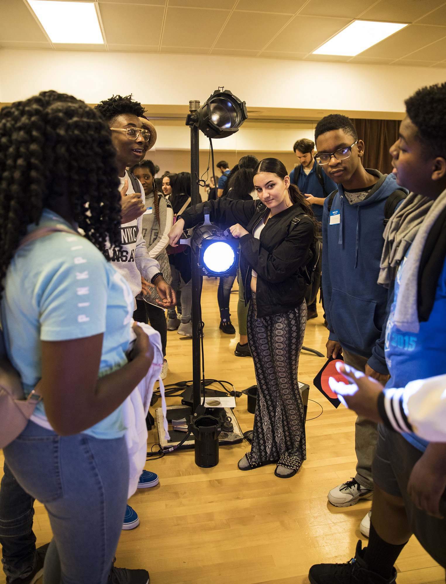 A group of five students, gather around a stage light setup in a school gymnasium. One female student points at the light while the others observe. They are participating in what appears to be a hands-on learning activity, reminiscent of enriching field trips in Kansas City.