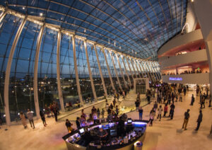 A large atrium with a curved glass ceiling at twilight, bustling with people. A bar area is in the foreground, and upper floors with balconies overlook the scene.