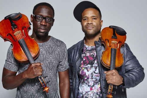 Two men holding violins. The person on the right is wearing a cap backward and a leather jacket; the person on the left wears glasses and a patterned shirt.