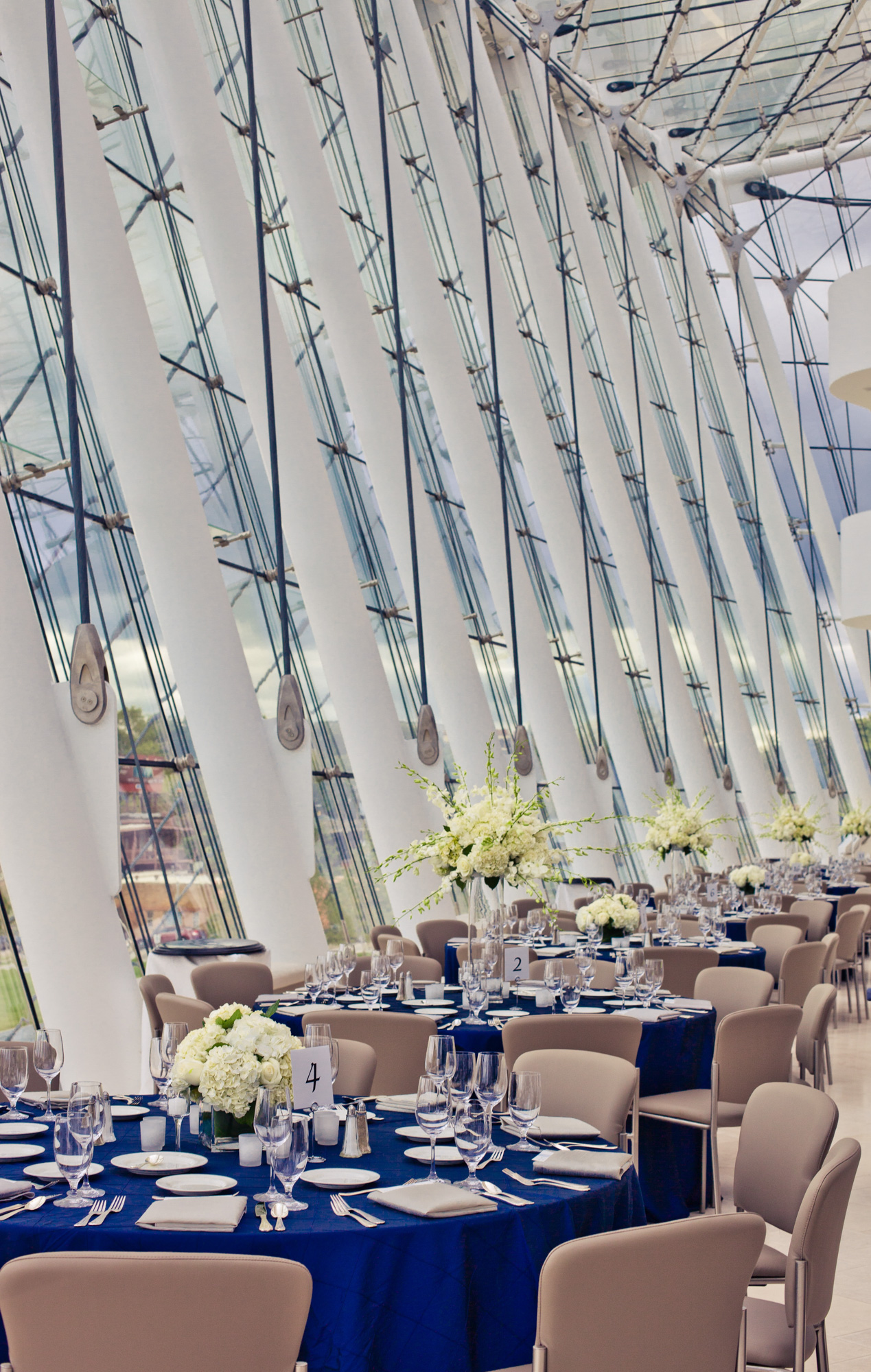 A modern banquet hall with tall, slanted windows, decorated with round tables covered in blue tablecloths, set with elegant tableware and white floral centerpieces, ideal for your next special event. Don't forget to fill out the Special Event Rental Form to secure your reservation.