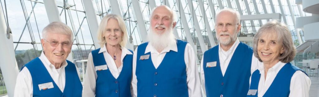 Five people wearing matching blue vests stand together indoors, smiling at the camera against a backdrop of large windows and modern architecture.