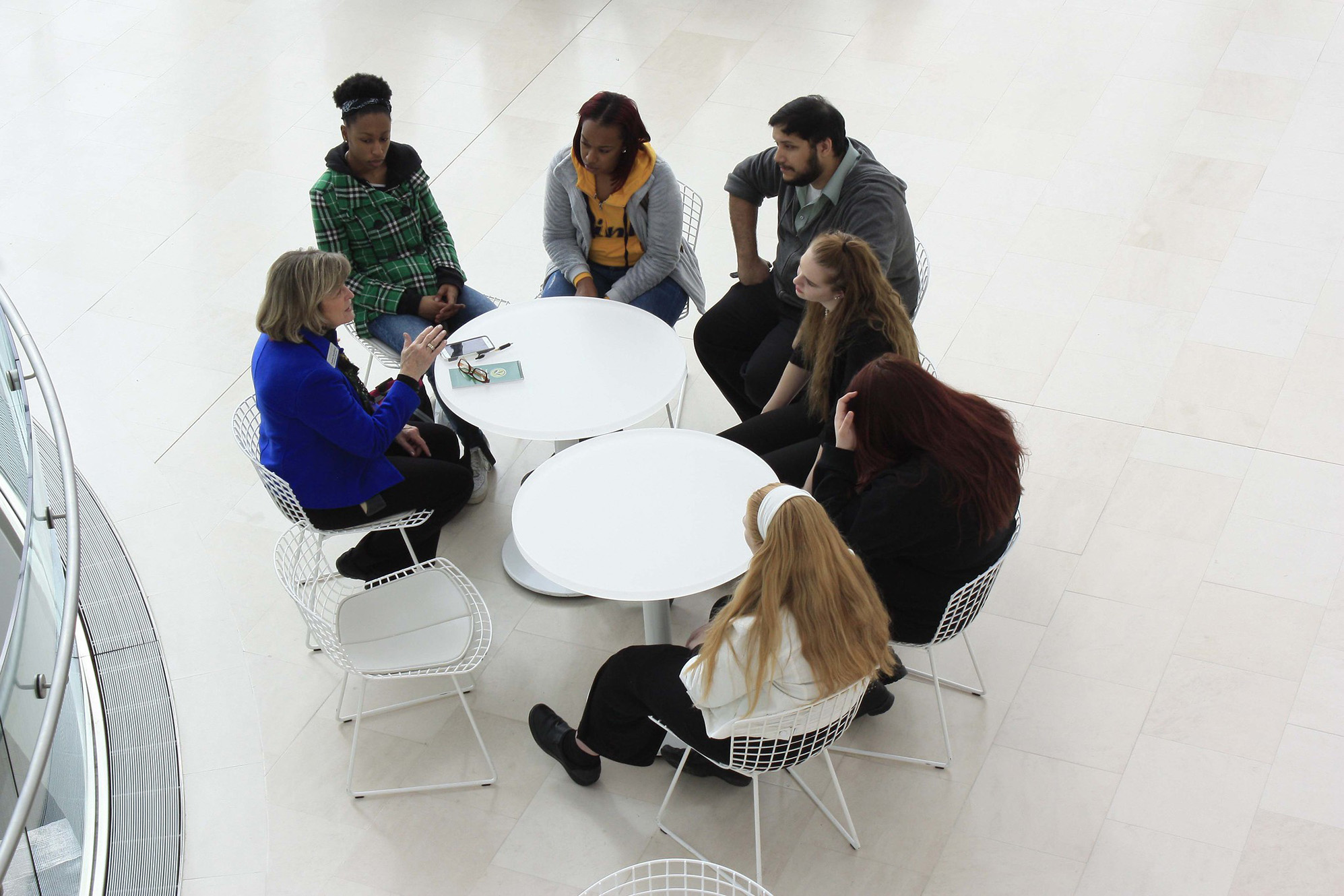 A group of six people, are seated around two circular white tables engaged in discussion. The setting appears to be a spacious, well-lit indoor area with light-colored flooring. One person, in a blue jacket, seems to be leading the conversation about field trips in Kansas City.