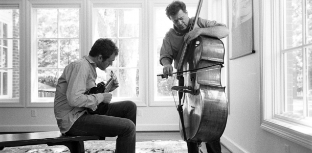 A man sitting and playing a mandolin while another man stands and plays a double bass in a well-lit room with large windows.