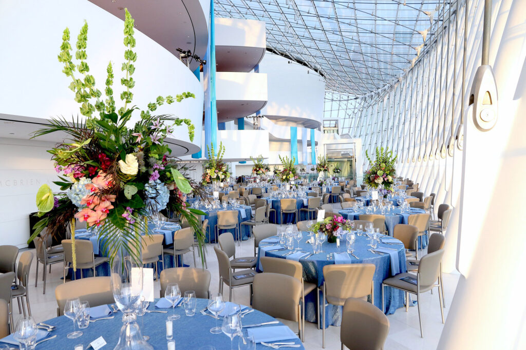 A spacious event hall with large windows, decorated with round tables covered in blue linens, elegant floral centerpieces, and neatly arranged table settings. Perfect for any occasion, you can now rent this exquisite space at the Kauffman Center.