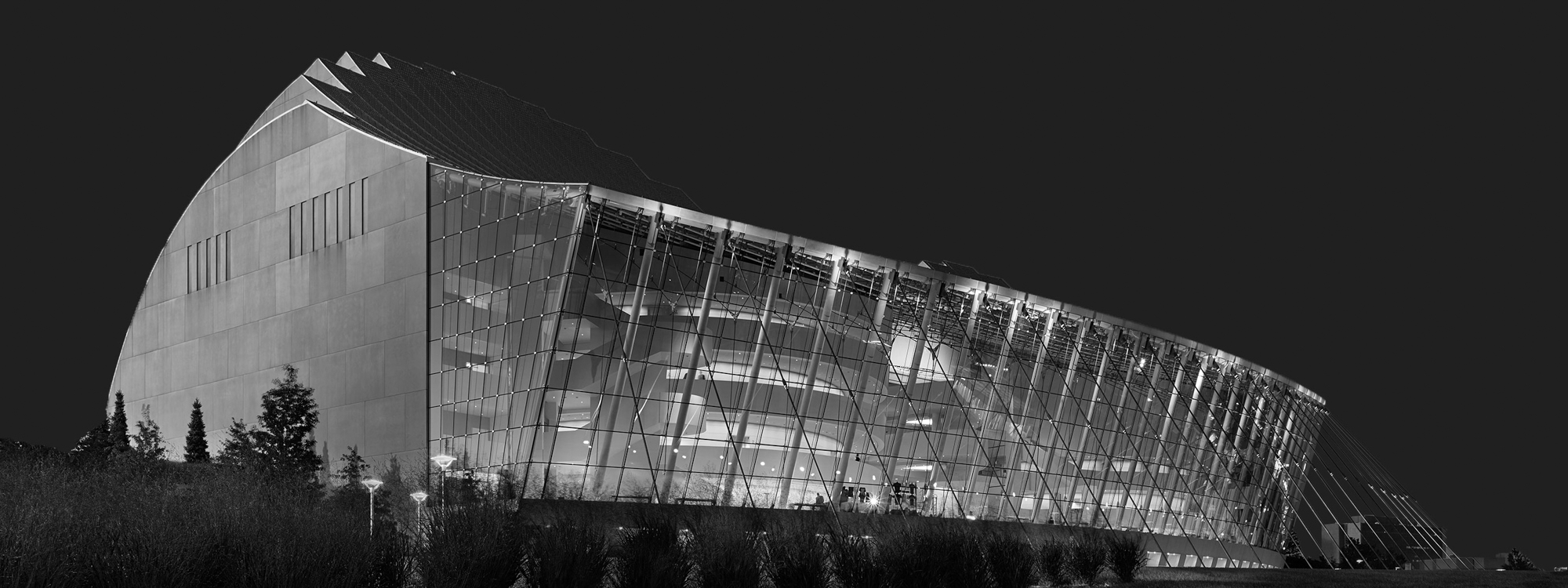 Black and white. Modern architectural building with a curved glass facade and illuminated interior, captured at night against a dark sky, showcasing innovative design in Architecture and Spaces.