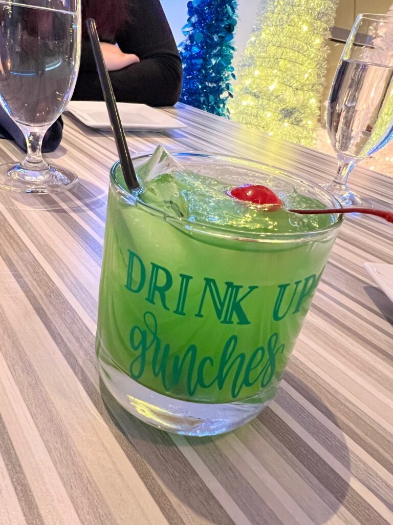 A green cocktail with a cherry garnish in a glass with "DRINK UP GRINCHES" text, placed on a striped tablecloth with water glasses and colorful decoration in the background.