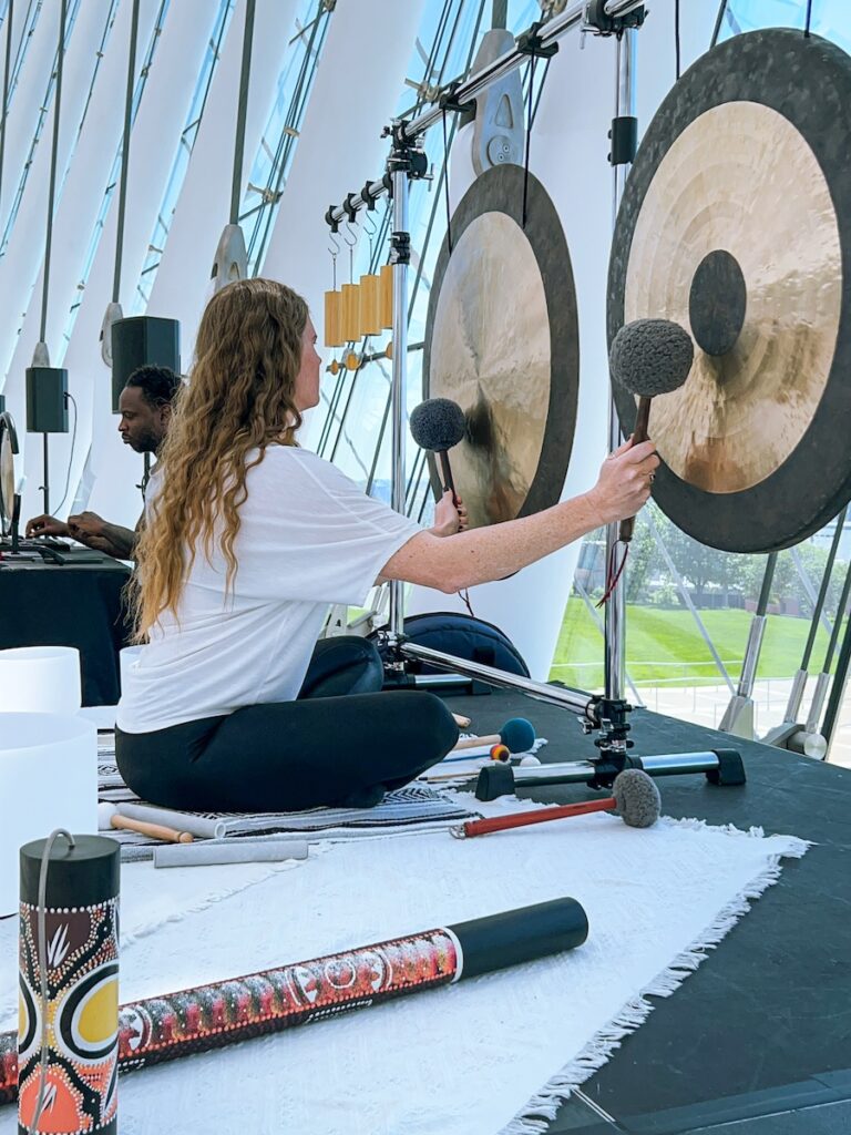 A woman sits cross-legged striking two large gongs with mallets during a sound bath at Kauffman Center, while a man in the background operates sound equipment. Various percussion instruments are arranged on a white mat.