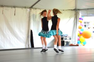 Two young girls in matching costumes perform a dance routine on a raised stage inside a tent, with colorful balloons in the background.