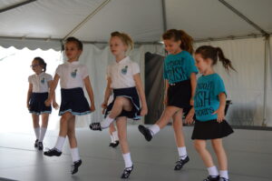 Five young girls are performing an Irish dance on a stage under a tent. Four girls wear matching white tops and navy skirts while the fifth wears a blue t-shirt and black skirt.