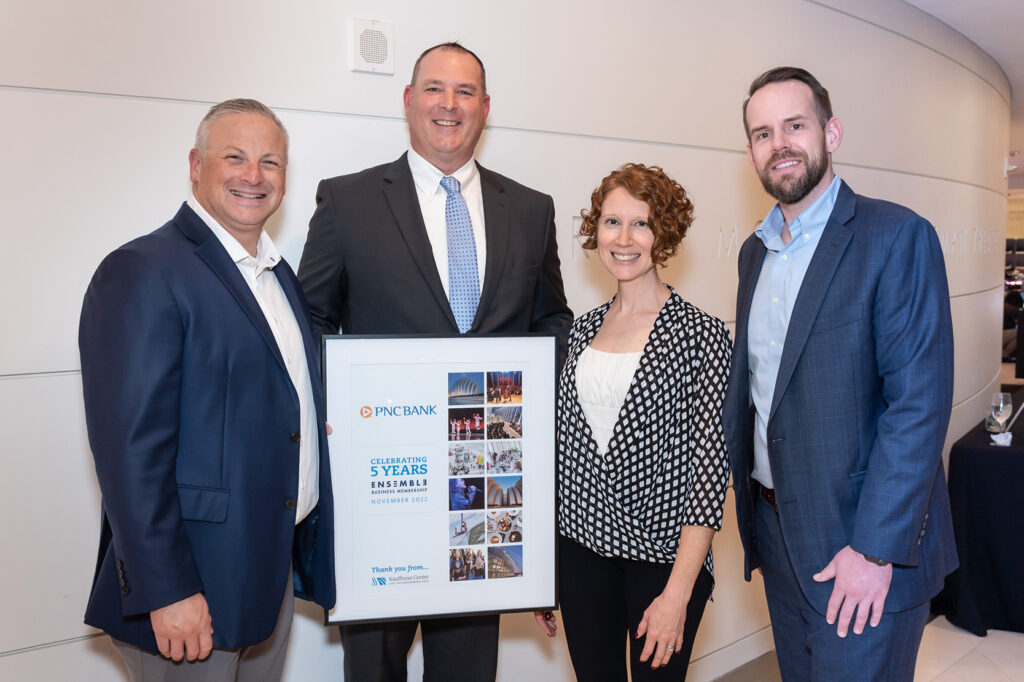 Four people stand together; one man holds a framed 5-year anniversary certificate from PNC Bank. Dressed in business attire and smiling, they proudly celebrate their business memberships and professional achievements.