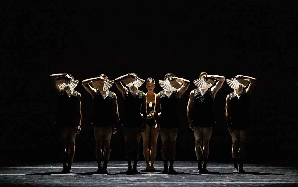 Six dancers in black attire perform on stage, holding white fans that partially obscure their faces, set against a dark background. This captivating Performing Arts Sampler showcases their graceful synchronization and expressive storytelling.