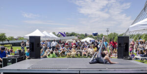 A performer balances a ball with their feet on stage at an outdoor event. The audience sits and stands under tents in the background on a sunny day.