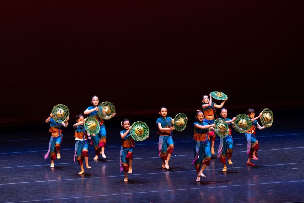 A group of children perform a dance on stage, each holding a green woven basket. They wear colorful costumes and are mid-movement against a dark background.