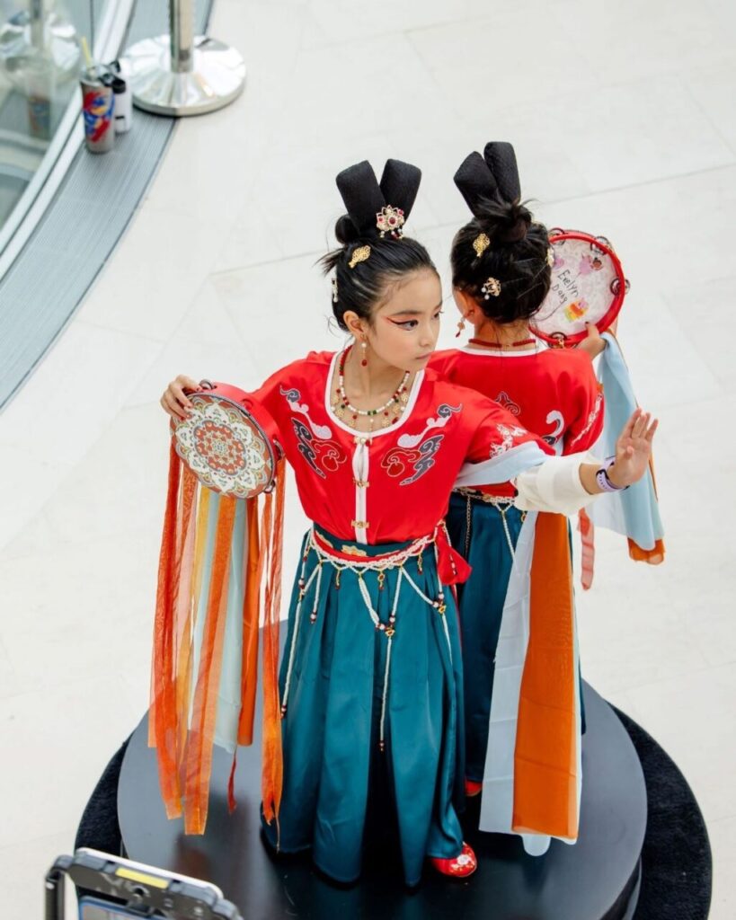 Two children in traditional Chinese attire stand back to back, each holding a decorated drum and ribbon.