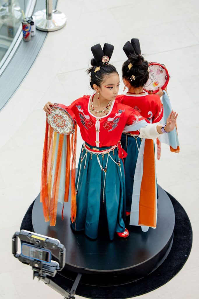 Two children in traditional Chinese attire stand back to back, each holding a decorated drum and ribbon.