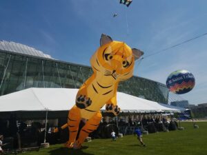 A large tiger-shaped kite is flying in front of a glass building with a white tent and crowd beneath it. A smaller kite and balloon with "Saint Luke's" written on it are also visible in the sky.
