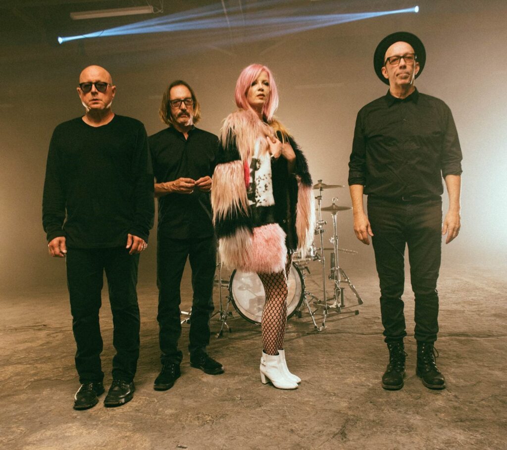 Four people are standing in front of a drum set. Three men wear black outfits, and a woman with pink hair wears a fur coat, fishnet stockings, and white boots.