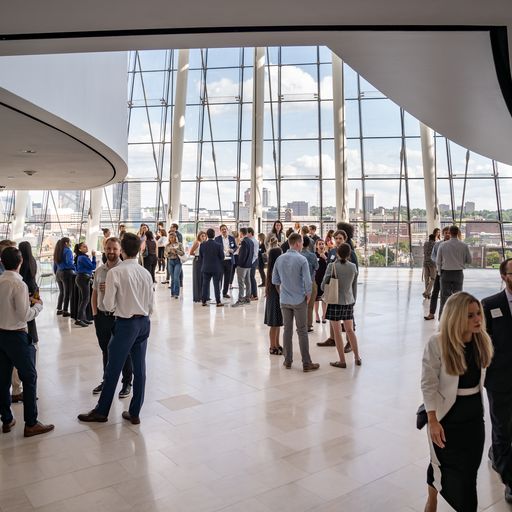 A group of people stand and converse in a spacious, modern building with large windows offering a city view in the background.