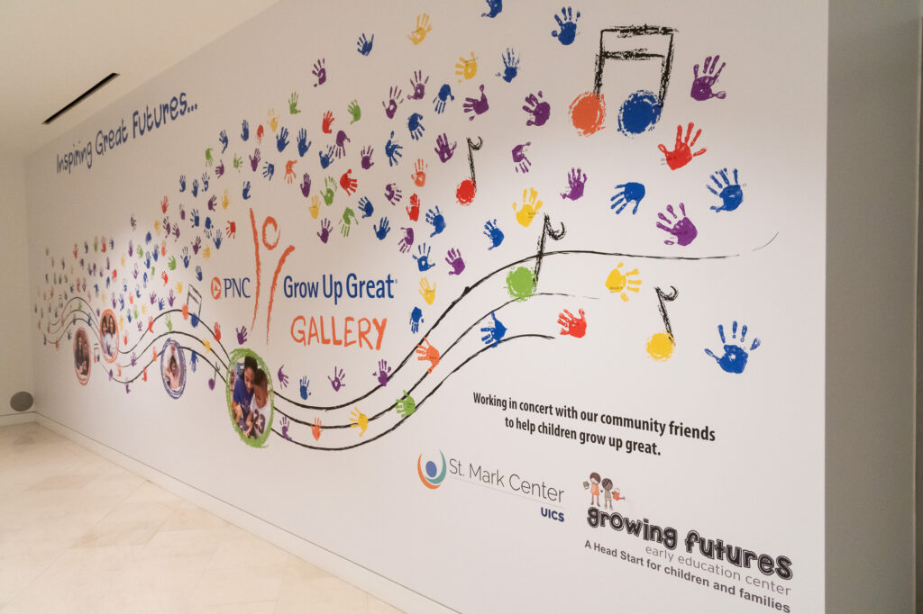 A wall mural with colorful handprints and logos for various children's programs, including PNC Grow Up Great Gallery and St. Mark Center, emphasizing collaboration to aid children's growth.