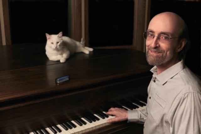 A man in glasses and a light-colored shirt plays a piano with a white cat lounging on top of it.