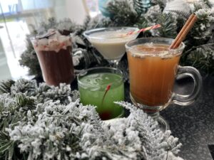 Four festive drinks placed on a frosted wreath: a hot chocolate garnished with marshmallows, a creamy cocktail in a martini glass, a green drink with a cherry, and a cinnamon-spiced drink.