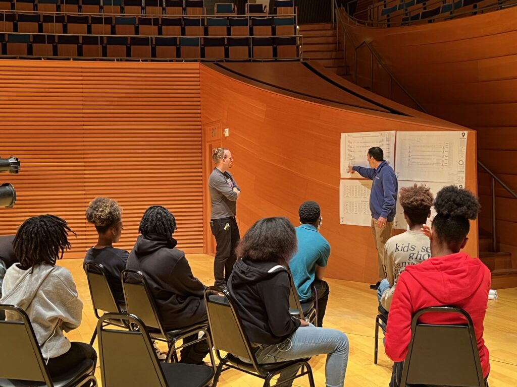 A group of students seated in chairs in an auditorium listen to two instructors in front of them, one pointing at a large piece of paper on the wall.