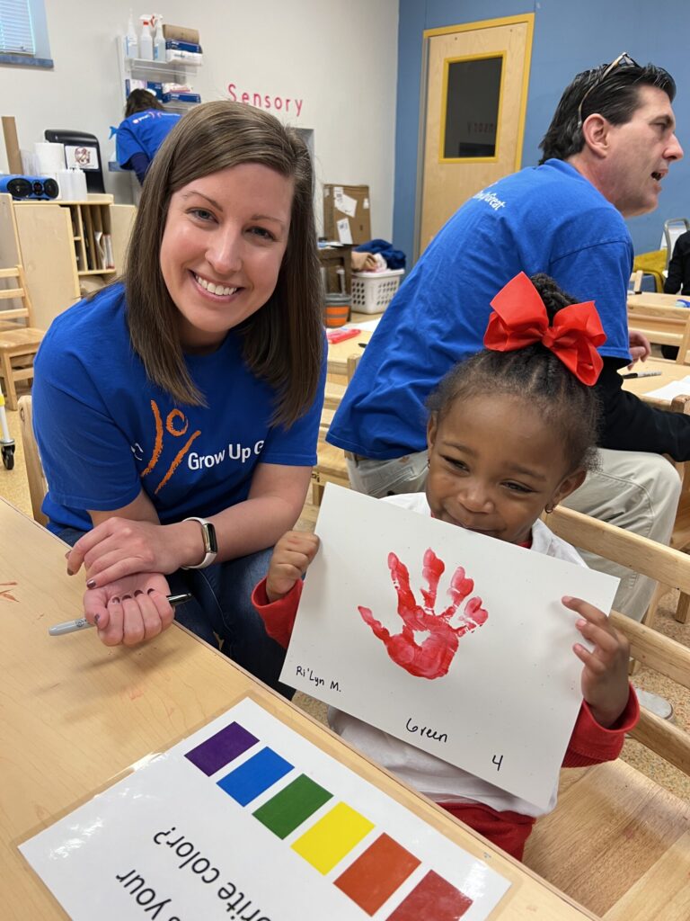 A woman and a young girl with a red bow in her hair are in a classroom. The girl is holding a paper with a red handprint.