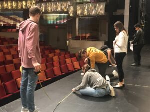Several people are working at the edge of a theater stage, measuring and marking positions on the floor.