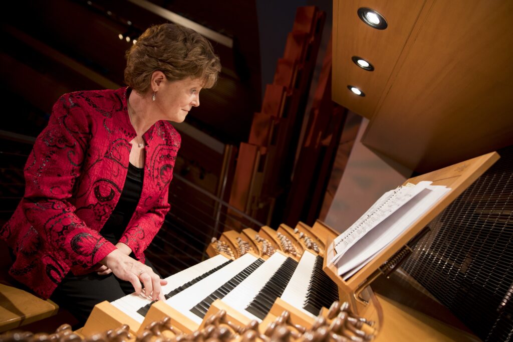 A person in a red jacket plays a large pipe organ, reading sheet music under overhead lights.