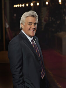 A smiling person with gray hair, dressed in a dark suit and striped tie, standing against a dimly lit background.